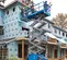 Side view of extended blue Genie scissor lift with two workers on platform, elevated in front of a house under construction.