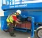 Side view of worker inspecting a blue Genie scissor lift with underneath compartment open.