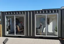 A modern container-style modular building with large glass windows, showing people seated inside—likely a meeting, consultation, or display space—while an elderly woman sits in a separate section on the right, suggesting a comfortable, residential or care-oriented interior.